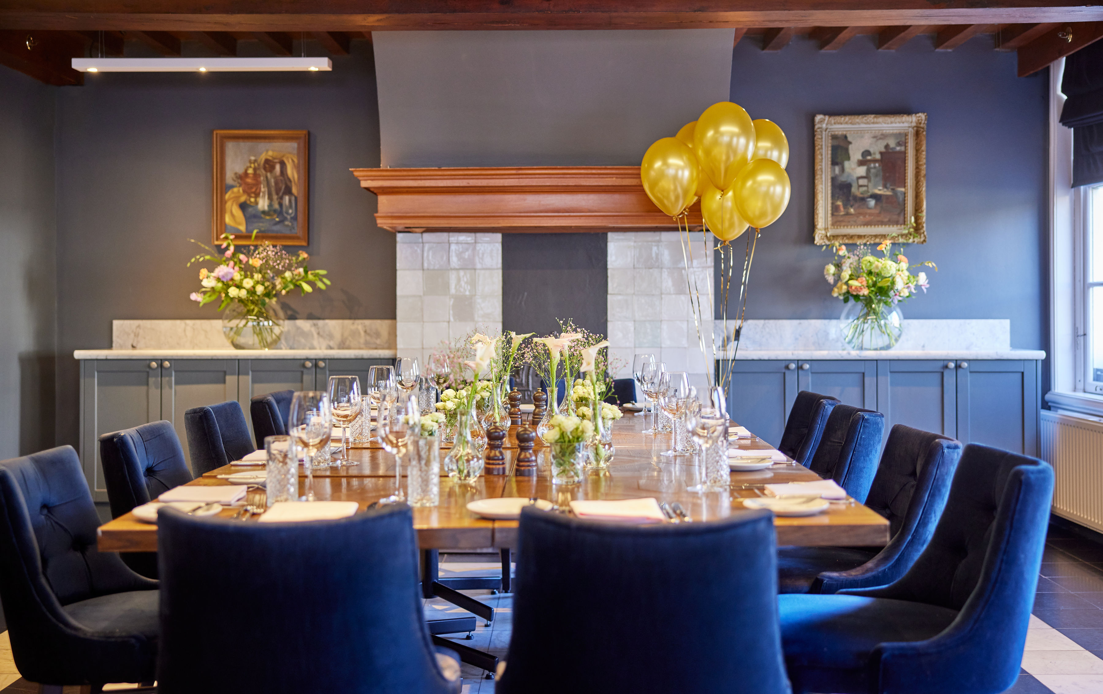A large dining table at the Copper Chamber at Pulitzer Amsterdam with flowers, blue chairs and balloons over a fireplace.