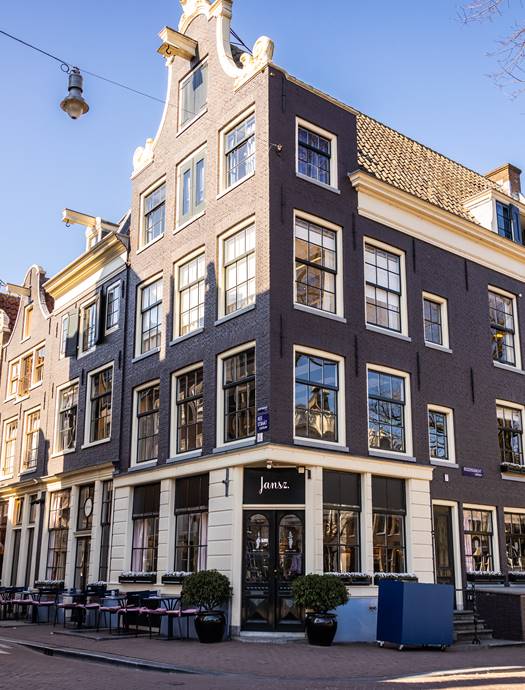 A corner building in Amsterdam with the Jansz. restaurant’s signage and outdoor seating area with a blue sky.