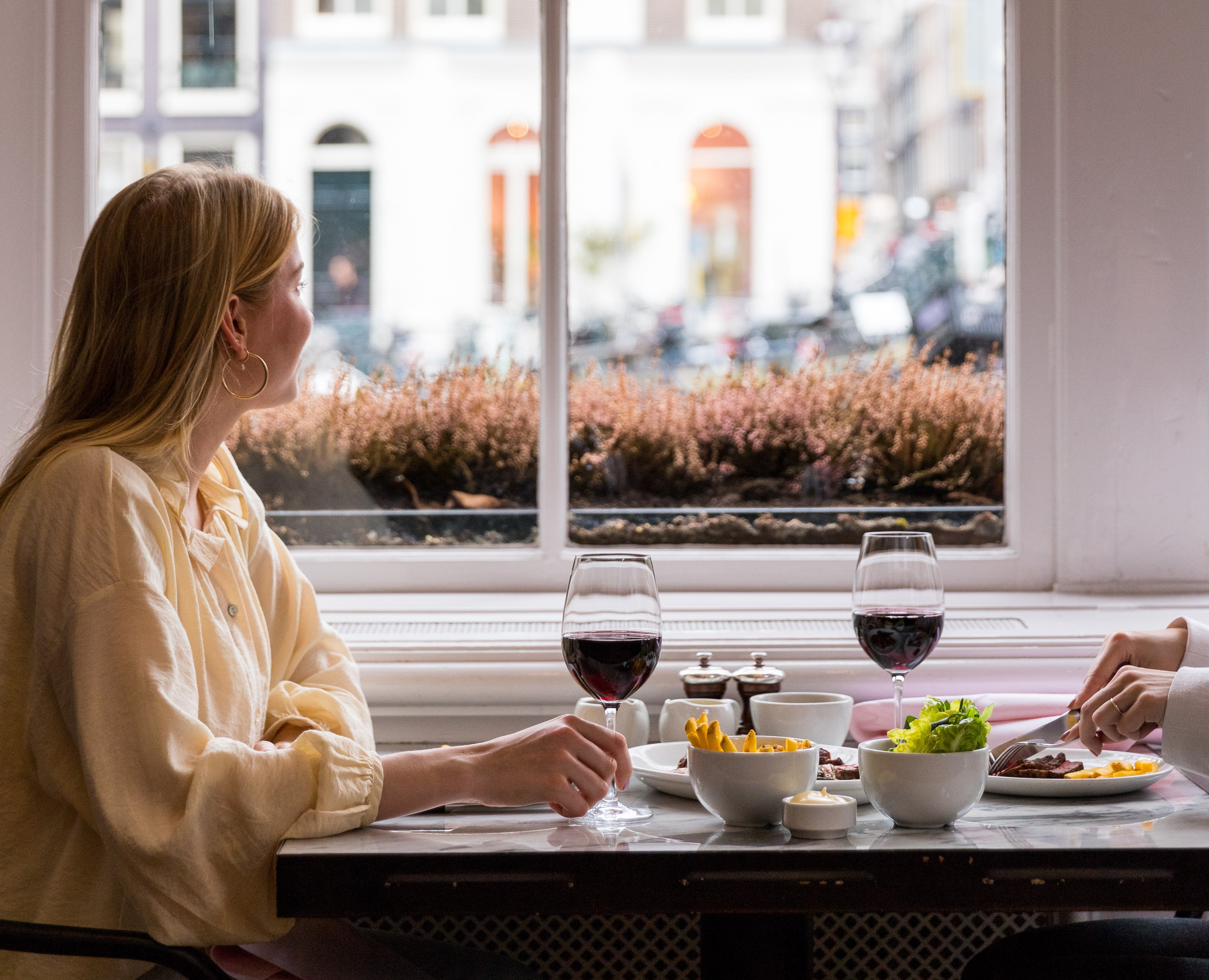 A window-side table for two at Jansz. with wine glasses and a woman enjoying the view of the streets of Amsterdam.